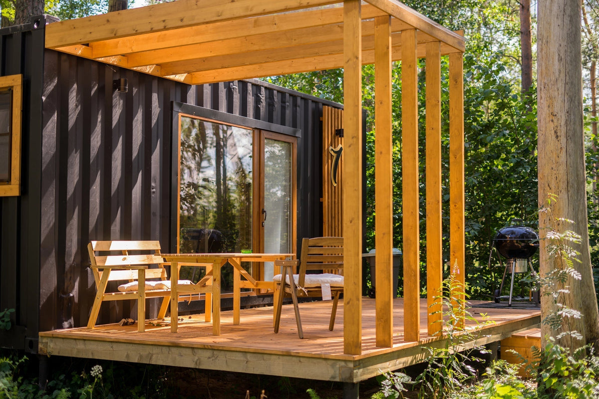Grillunterstand selber bauen – ein überdachter Holzunterstand auf einer Terrasse mit Bank und Tisch vor einem Containerhaus.